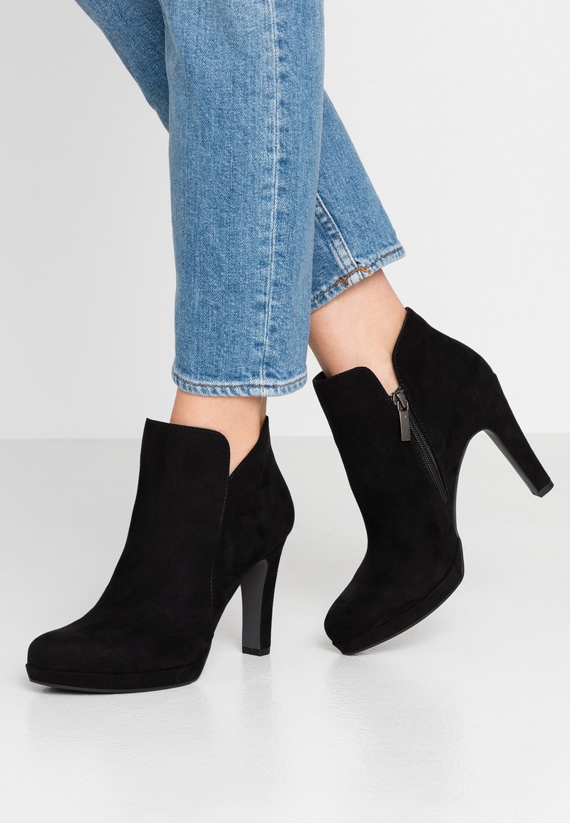 Jambes en jean bleu portant des bottines à talons hauts en suède noir avec fermetures éclair latérales, posées sur un sol et un fond clairs.