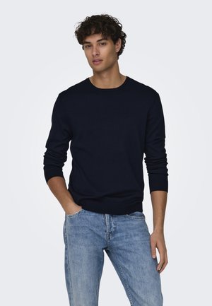 Jersey de punto azul marino con escote redondo y mangas largas, combinado con unos vaqueros de denim azul claro. El jersey tiene un dobladillo acanalado ajustado.