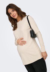 Έγκυος γυναίκα που φορ άει μπεζ μπλούζα με μακριά μανίκια και κουμπιά στο πλάι, μαύρο παντελόνι και μικρή μαύρη τσάντα ώμου.