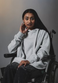 Grå hettegenser med avslappet passform, båret over svarte glitrende bukser. Modellen sitter i rullestol, med langt mørkt hår og øreringer.