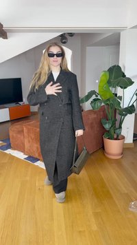 Schwarz doppelt geknöpfter Mantel aus strukturiertem Stoff, getragen über einem weißen Oberteil und schwarzen Hosen. Passende graue Stiefel und eine dunkle Handtasche.