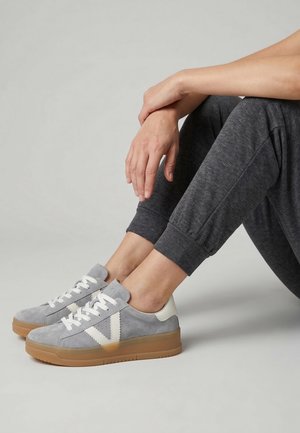 Person sidder med bøjede ben iført grå joggingbukser og grå ruskindssneakers med hvide snørebånd og gummisåler mod en neutral baggrund.