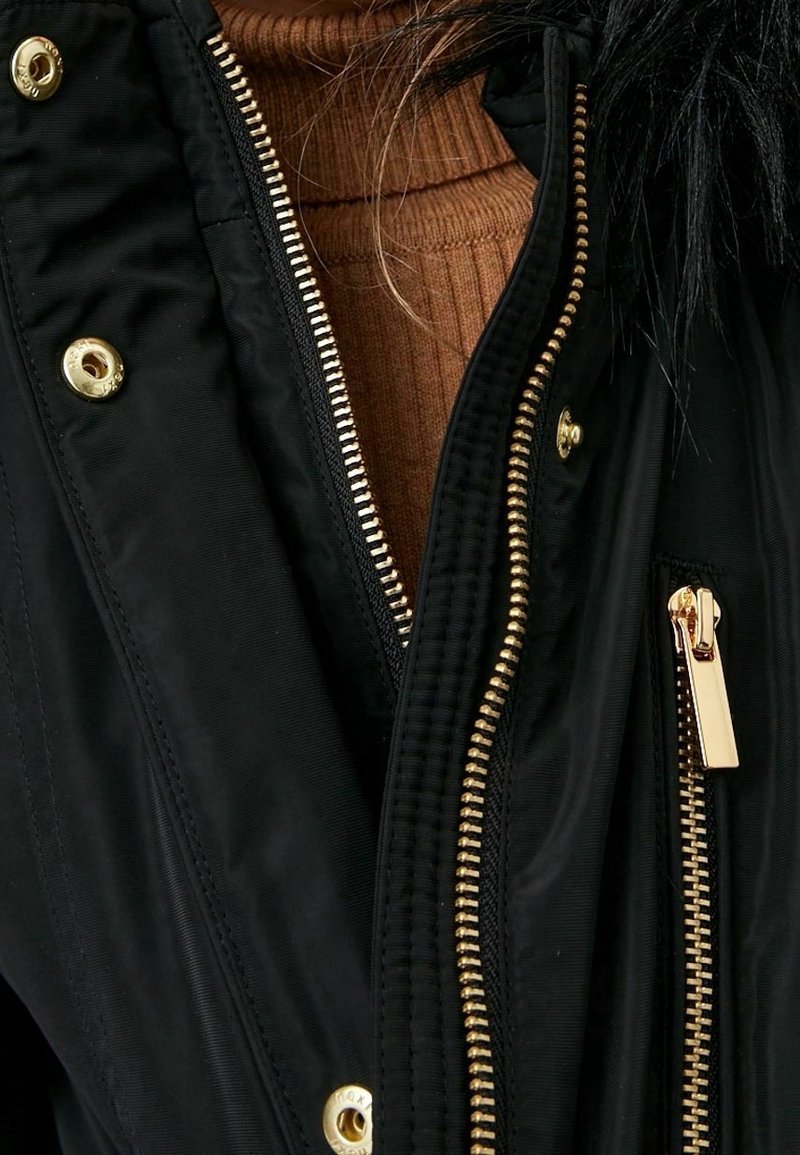 La veste noire présente des fermetures éclair dorées, des boutons pression et des accents en peluche. L'intérieur révèle un pull côtelé beige en dessous. Tissu doux, coupe ajustée.