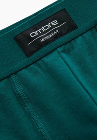 Sous-vêtements pour homme en bleu sarcelle avec une ceinture douce, arborant une étiquette noire avec "Ombre MENSWEAR." Texture lisse et détails cousus.