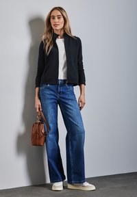 Giacca nera trapuntata con zip, t-shirt bianca, jeans blu a gamba larga, sneakers crema e una piccola borsa marrone. In piedi contro un muro grigio.