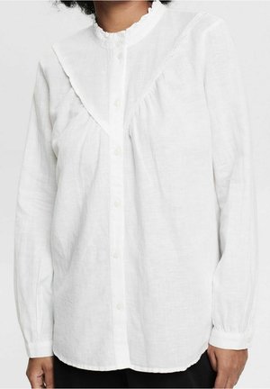Blouse blanche à manches longues boutonnée, avec col à volants et empiècement, portée par une personne ayant des cheveux bouclés foncés visibles sur le bord supérieur.