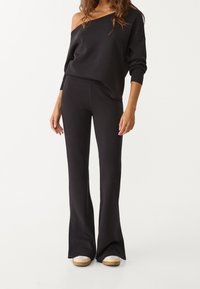 Sudadera negra con hombros descubiertos y mangas largas, combinada con pantalones acampanados negros. Ambas prendas están hechas de una tela suave y texturizada. Calzado casual incluido.