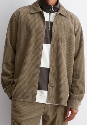 Mężczyzna ubrany w brązową kurtkę sztruksową na czarno-białym swetrze z suwakiem do ćwiartki, w połączeniu z dopasowanymi brązowymi sztruksowymi spodniami, ręka w kieszeni.