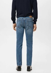 Mężczyzna ubrany w dopasowane niebieskie dżinsy, czarny sweter i czarne buty, stojący tyłem na prostym białym tle.