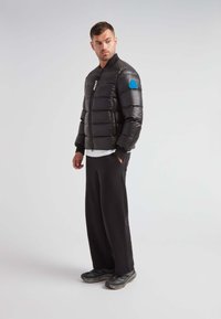 Giacca nera imbottita con zip frontale, polsini a coste e una patch blu con logo sulla spalla. Indossata con pantaloni neri e scarpe sportive.