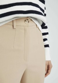 Pantalons beiges ajustés avec une texture subtile, dotés d'une fermeture à bouton et de poches latérales. Un pull rayé noir et blanc couvre partiellement la taille.