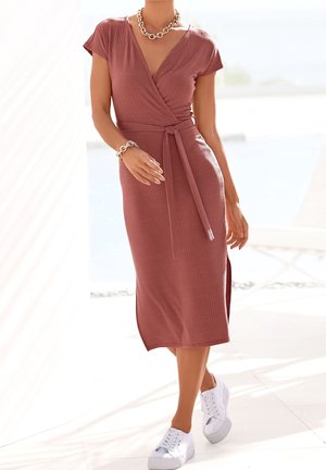 Femme portant une robe midi portefeuille mauve à manches courtes, nouée à la taille, des baskets blanches, ainsi qu'un collier et un bracelet chaîne en or massif, debout à l'intérieur.