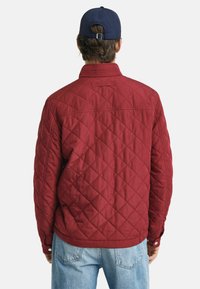 Rode gewatteerde jas met een opstaande kraag, elastische manchetten en een tailleband. De stof heeft een ruitpatroon. Gekozen in combinatie met lichtblauwe jeans.