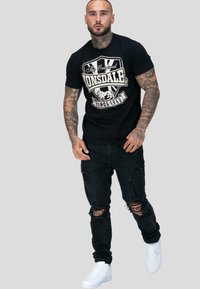 Mies, jolla on tatuointeja, pukeutuneena mustaan Lonsdale-paitaan, revittyihin mustiin farkkuihin ja valkoisiin tennareihin, seisoo vaaleaa taustaa vasten.