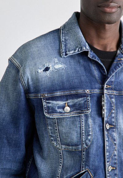 Denim jakna u plavoj boji s istrošenim detaljima, uključujući poderani komad na prsima. Sadrži metalne dugmadi i dva prednja džepa.