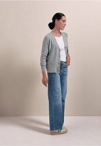 Grau gestrickte Strickjacke mit vorderen Knöpfen, getragen über einem weißen Oberteil und hoch taillierten hellblauen Jeans. Legere Schuhe runden den Look ab.