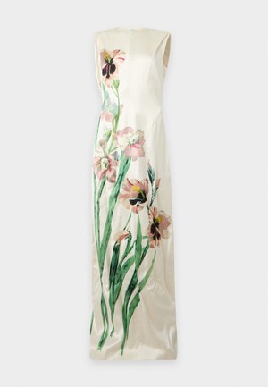 Femme portant une robe blanche sans manches longue jusqu'au sol avec un grand imprimé floral rose et vert, debout les mains derrière le dos.