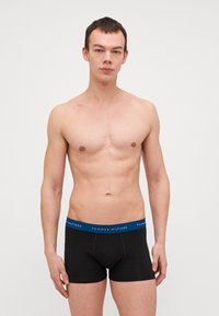 Musta bokserihousut sinisellä vyötärönauha, jossa on "TOMMY HILFIGER" -logo. Kangas näyttää sileältä, istuva muotoilu lantion ympärillä.