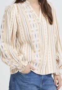 Blouse à manches longues avec un col en V, présentant un motif géométrique jaune, blanc et noir, et des poignets froncés. Tissu doux et léger.