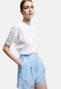 Hvid bomulds T-shirt med broderede øje-udskæringer ved halsen, parret med lyseblå shorts med bølgede kanter og blomsterudskæringer.