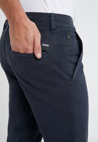 Tmavě modré kalhoty z hladké látky se slim fit střihem. Obsahují kovovou logo nášivku na zadní kapse a detail v oranžovém prošívání.