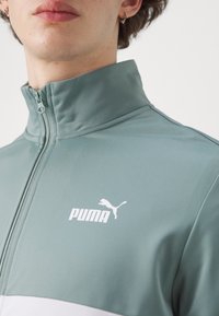 Grøn zip-up jakke med høj krave, der har hvidt logo og farveblokeringsdesign. Blødt stof med en glat tekstur.
