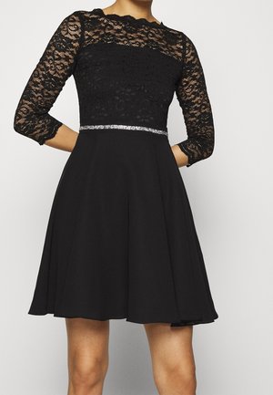 Femme portant une robe à manches trois-quarts en dentelle noire avec une ceinture argentée à la taille, debout les mains derrière le dos.