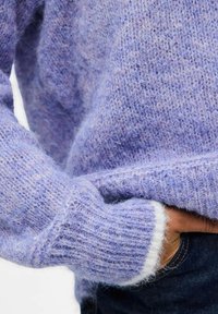Main dans un jean foncé, posée dans la poche, couverte par un pull en tricot lavande duveteux avec un détail de poignet blanc.