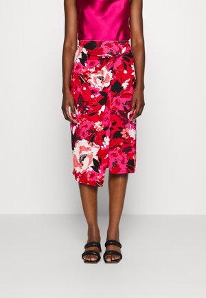 Kvinde iført en skinnende pink satin top, rød blomsterprintet omslagsnederdel og sorte sandaler med dobbelt rem, stående mod en ensfarvet lys baggrund.