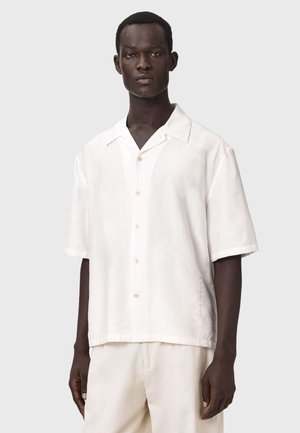 Ung mand med mørk hud iført en løs, hvid kortærmet skjorte med knapper og hvide bukser, stående foran en lysegrå baggrund.