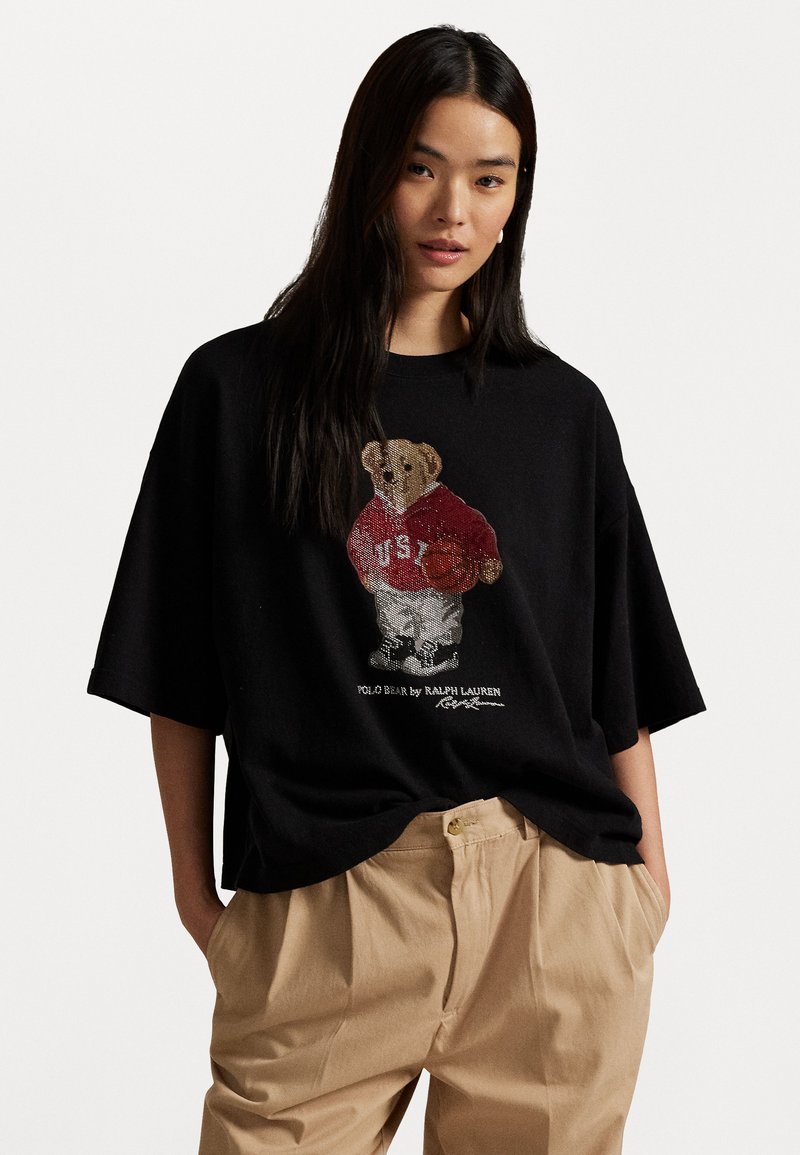 Črna oversize majica s kratkimi rokavi, ki prikazuje grafiko medveda v rdečem dresu z košarko, na enostavni beli podlagi.