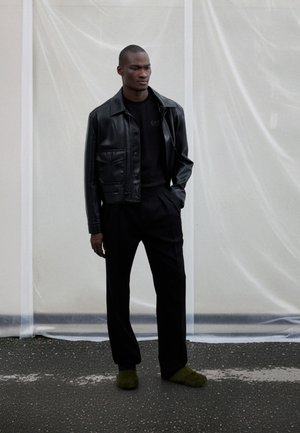 Černá kožená bunda s předními kapsami, nošená přes černé tričko. Kombinováno s černými kalhotami na míru a zelenými chlupatými nazouvacími botami.