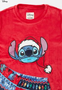 Czerwona bluza z polaru z naszytym wizerunkiem postaci Stitcha w czapce świętego Mikołaja, zestawiona z wzorzystymi spodenkami do piżamy w niebieskich i świątecznych kolorach.