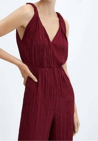 Combinaison plissée bordeaux avec un décolleté en V, des bretelles larges et une taille ajustée. Fabriquée en tissu texturé léger, avec des poches latérales.