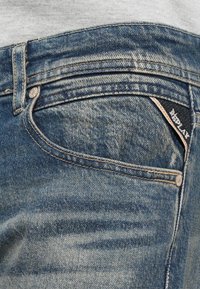 Jeans de ganga azuis com uma textura desbotada e desgastada, apresentando um bolso para moedas e um rótulo branco no bolso de trás.