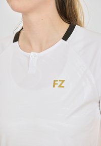 Valkoinen urheilupaita, jossa on pieni kulta "FZ" -logo, nappilistat ja musta kaula-aukko. Sileä rakenne, jossa on hienovarainen kuvio.