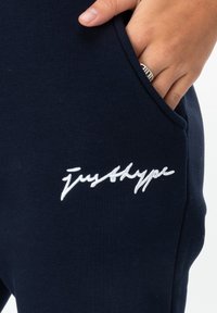 Navy blue joggers featuring a white embroidered logo "justhype." Soft fabric with side pockets, fitted silhouette, and minimalistic design.