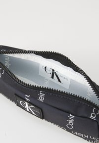 Pochette nera con zip caratterizzata da un motivo a ripetizione del logo "Calvin Klein Jeans". All'interno presenta una fodera chiara con un'etichetta del marchio.