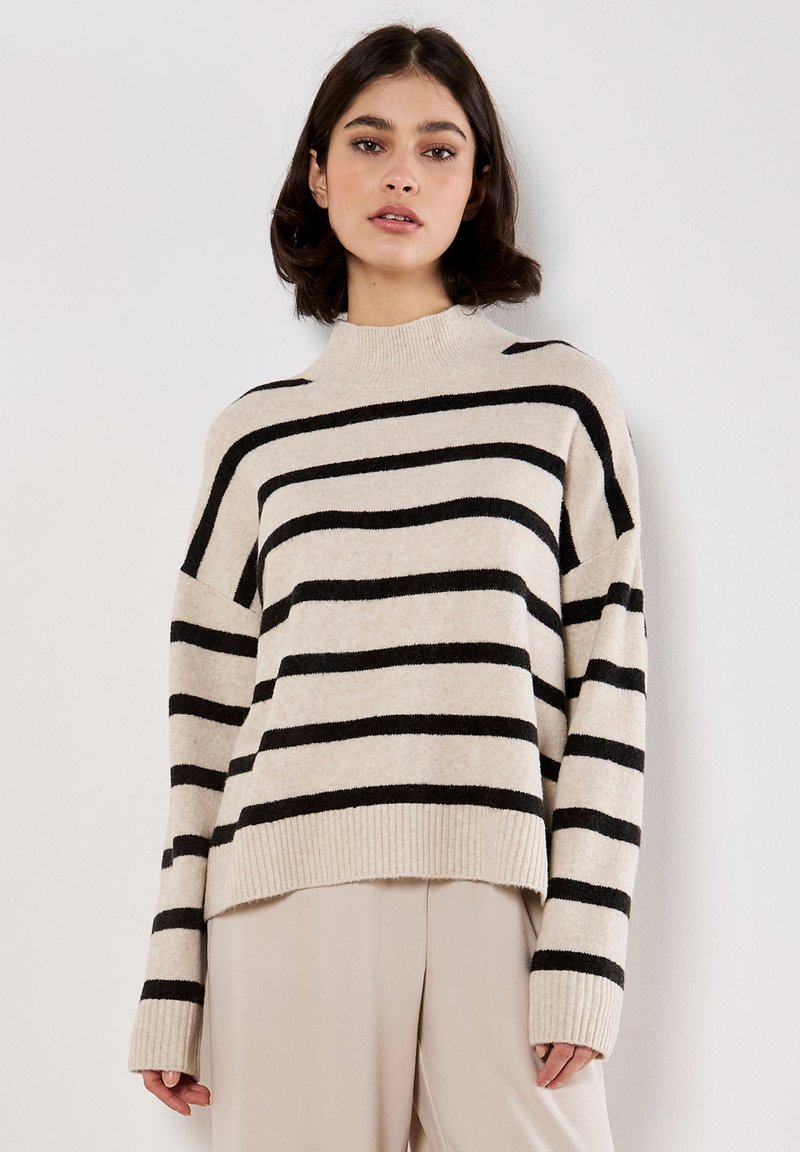 Maglione beige a righe con strisce orizzontali nere, scollatura a coste e polsini, vestibilità comoda, texture morbida. Abbinato a pantaloni di colore chiaro.