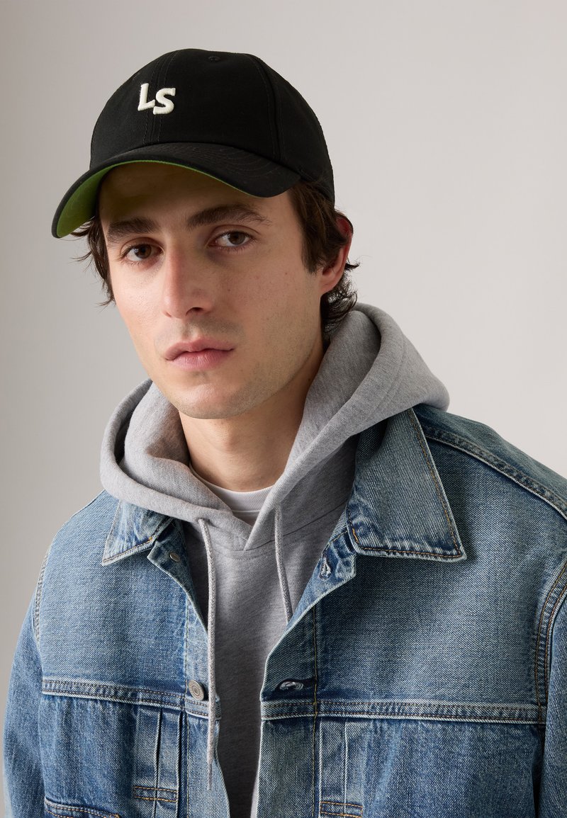 Chaqueta de mezclilla con lavado azul claro, sudadera gris debajo y gorra negra con logo blanco. Presenta un collar clásico y botones frontales.