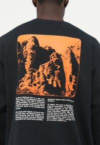 Melns sporta krekls ar oranžu grafiku, attēlojošu raupjas klints un baltu tekstu apakšā, kas apraksta Big Bend nacionālo parku.