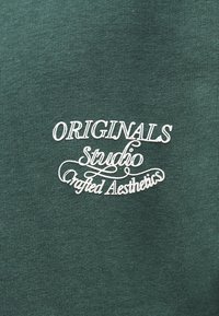Πράσινο ύφασμα με κεντημένα λευκά γράμματα: «ORIGINALS Studio Crafted Aesthetics.» Ομαλή υφή, κοντινή λήψη του σχεδίου.