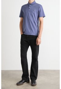 Fialové polo tričko s krátkým rukávem, límečkem a třemi knoflíky, v kombinaci s černými rovnými džíny a tmavými botami, nosí stojící postava.