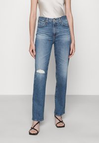 Vysoké modré džíny z denimu s poškozeným kolenem. Rovný střih v kombinaci s černými sandály na pásku, jemná textura látky.