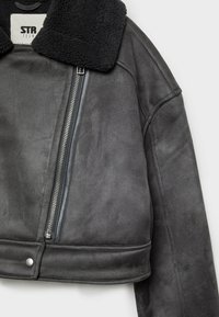Veste en daim gris foncé avec col en peau lainée noire, fermeture éclair avant asymétrique et bouton-pression au bas de l'ourlet sur fond blanc.