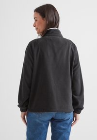 Black fleece jacket with a high collar, relaxed fit, and ribbed cuffs, paired with light blue denim jeans. Smooth texture, no visible patterns.