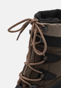 Botte en tissu marron avec un dessus noir, comportant un col en fourrure, des lacets contrastants et un matériel robuste pour un maintien sécurisé.