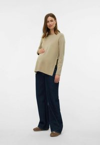 Top beige a maniche lunghe con fessure laterali, abbinato a jeans larghi in denim scuro, su uno sfondo bianco minimalista.