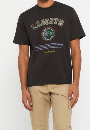 Homme portant un T-shirt noir Lacoste avec le logo Tennis Heritage, un texte et une signature, assorti à un pantalon beige.