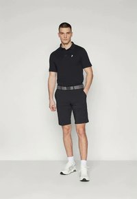 Noor mees seisab, üks käsi taskus, seljas must polo särk, mustad lühikesed püksid, hall vöö, valged sokid ja valged tossud.